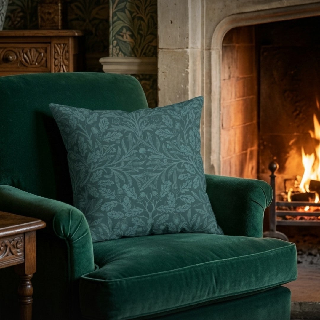 A square decorative throw pillow with a muted blue-green damask floral pattern, resting on a deep emerald green velvet armchair in front of a warm, glowing fireplace.