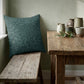 A square teal throw pillow with a subtle damask leaf pattern rests on a rustic wooden bench next to a matching wooden table decorated with ceramic vases and greenery.