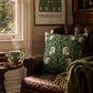 A decorative throw pillow with a green William Morris-style floral pattern sits on a brown leather armchair in a cozy room setting with a wooden side table, a steaming mug, and a framed William Morris print on the wall.