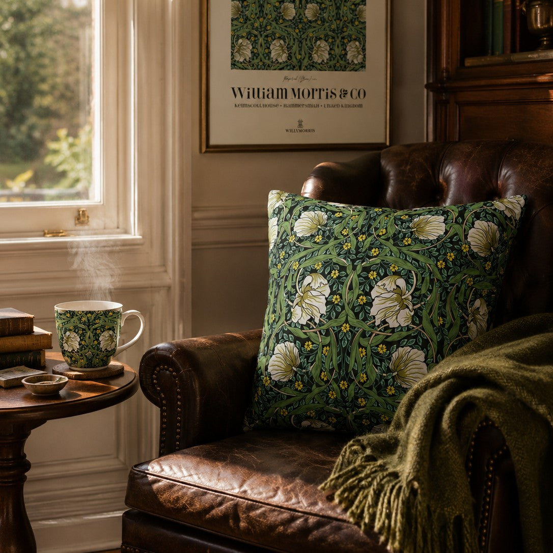 A decorative throw pillow with a green William Morris-style floral pattern sits on a brown leather armchair in a cozy room setting with a wooden side table, a steaming mug, and a framed William Morris print on the wall.