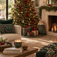 A cozy holiday living room scene with a decorated Christmas tree, wrapped gift boxes, patterned storage bags, candles, pillows, and a fireplace mantel with greenery.