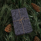 Gift wrapped in dark paper with a floral pattern, tied with string, on green foliage with pine cones.