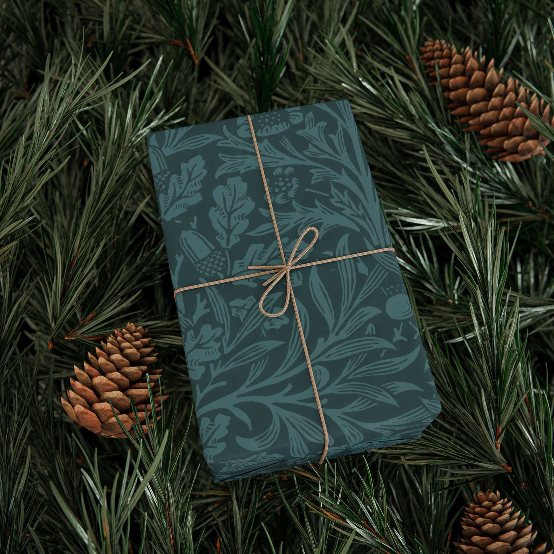 Gift wrapped in blue paper with a floral pattern, tied with brown string, on green foliage with pine cones.