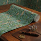 A roll of decorative wallpaper with a botanical pattern in mint green, olive green, and brown, partially unrolled on a wooden table next to a brown leather folio, a gold pen, and dried lavender flowers.