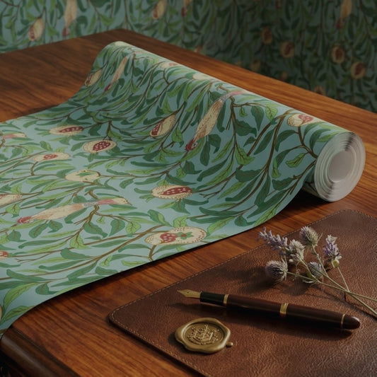 A roll of decorative wallpaper with a botanical pattern in mint green, olive green, and brown, partially unrolled on a wooden table next to a brown leather folio, a gold pen, and dried lavender flowers.