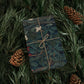 Gift wrapped in a dark floral pattern with brown string, surrounded by pine branches and cones.