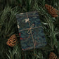 Gift wrapped in a dark green floral pattern with brown string, surrounded by pine branches and cones.