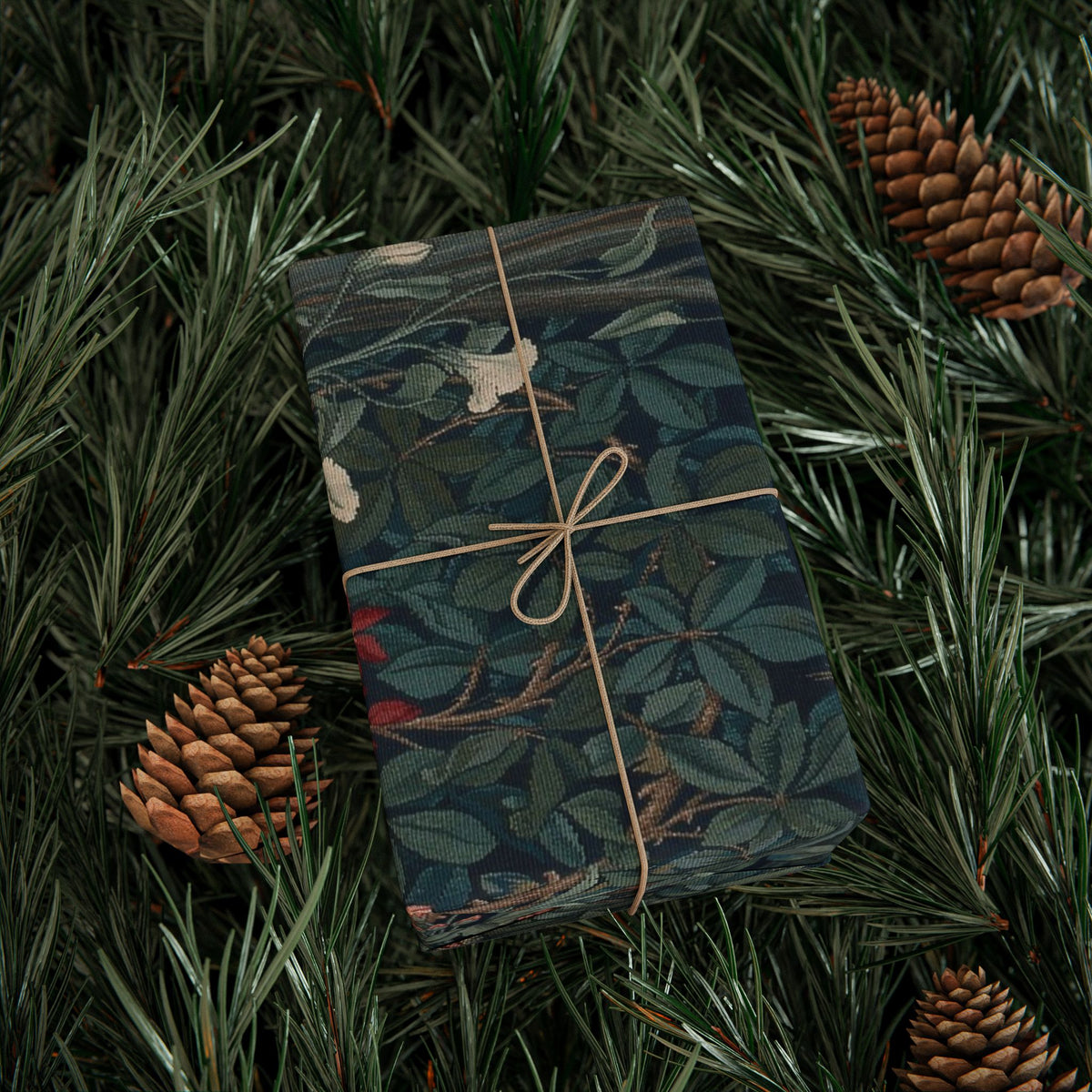 Gift wrapped in a dark floral pattern with brown string, surrounded by pine branches and cones.