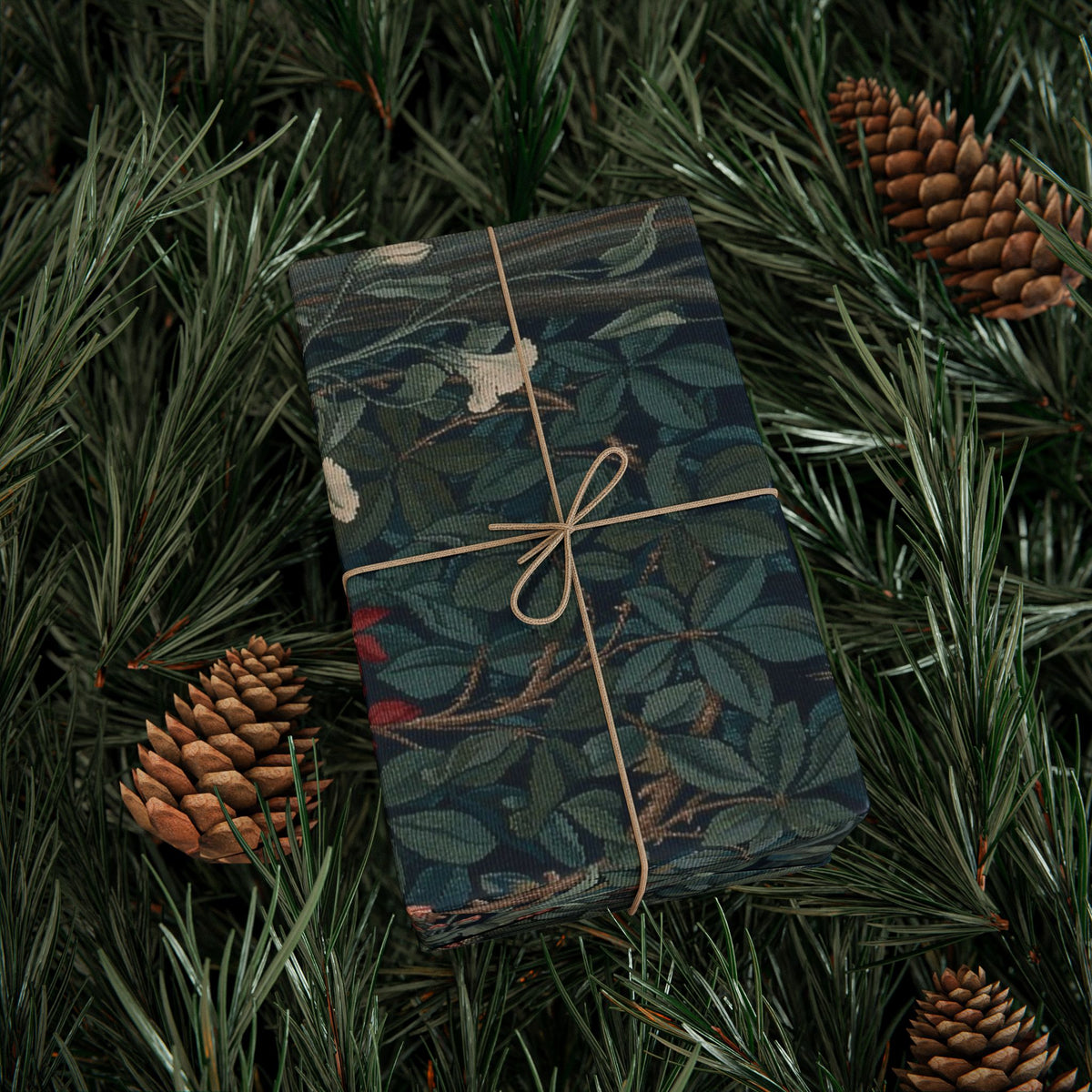 Gift wrapped in a dark green floral pattern with brown string, surrounded by pine branches and cones.