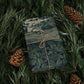 Gift wrapped in green floral paper with twine, surrounded by pine branches and cones.