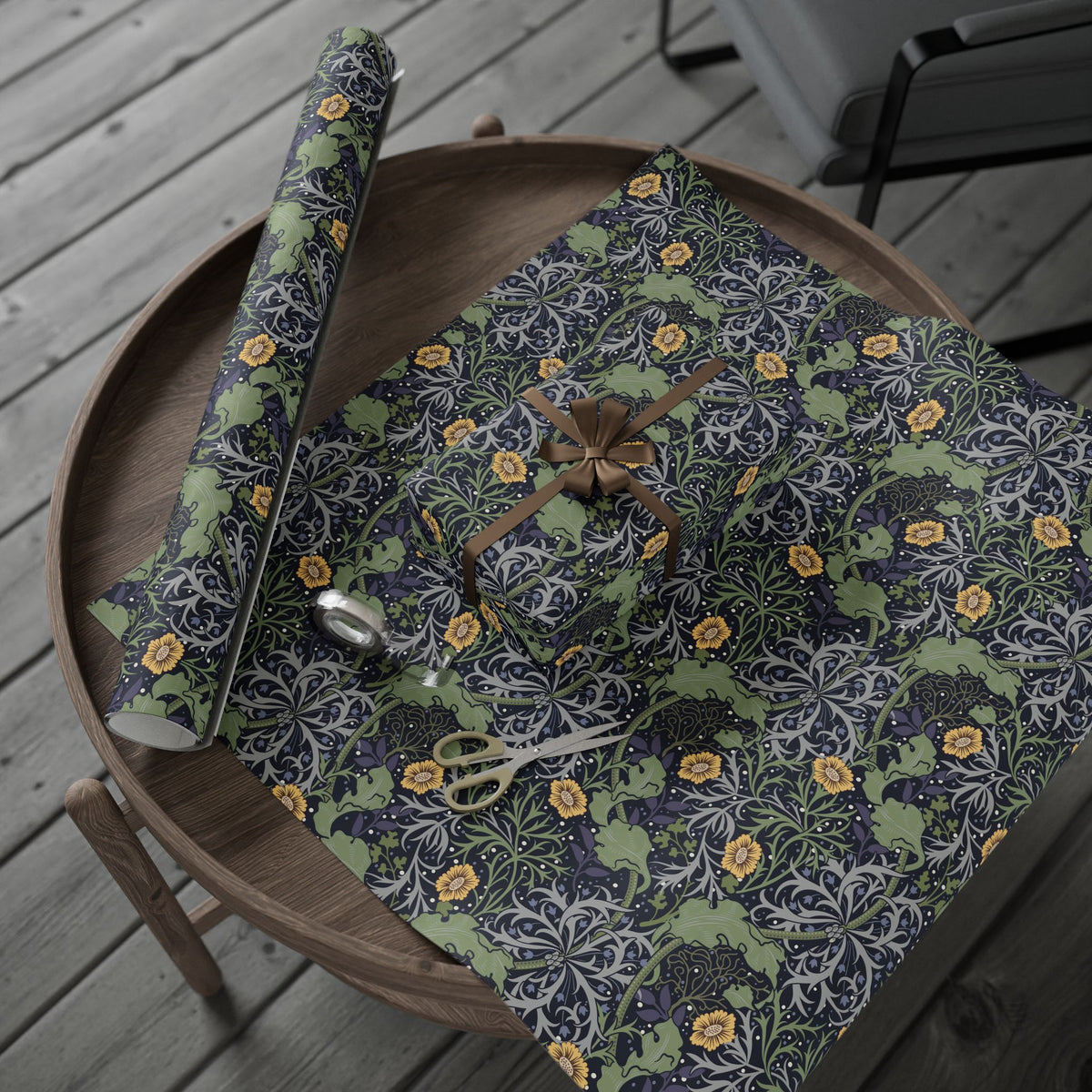 Round wooden table with a floral-patterned cloth, scissors, and a roll of green paper on a wooden deck.