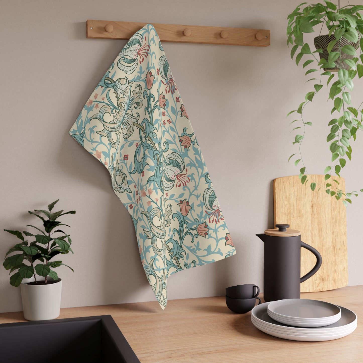 A kitchen towel with a William Morris-style floral and scroll pattern in teal, rust, and cream hangs from a wooden peg on a beige wall above a kitchen counter with dishes and plants.