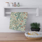 A patterned kitchen towel with a teal and rust botanical design on a cream background, hanging from a white wall-mounted rack in a bright kitchen setting.