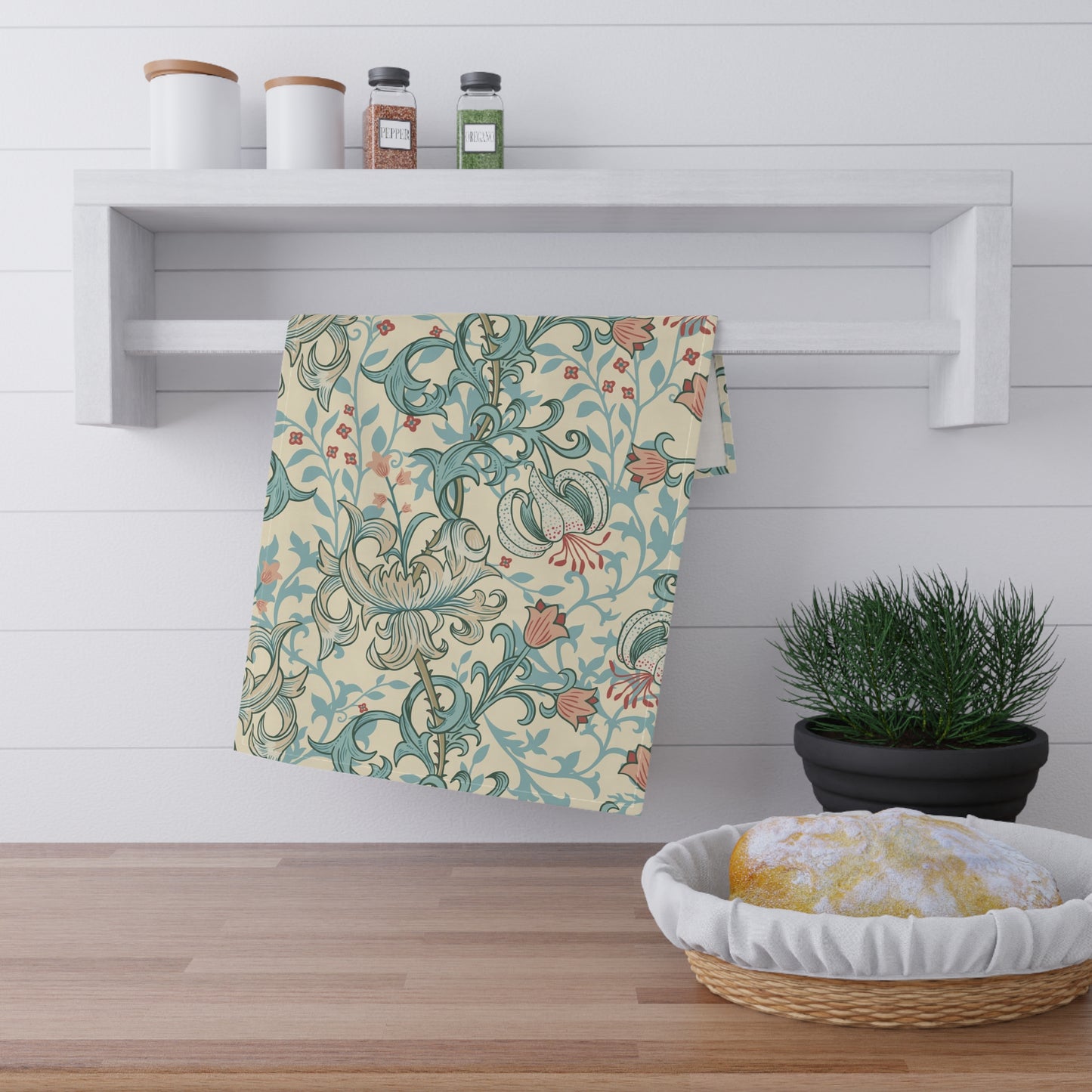 A patterned kitchen towel with a teal and rust botanical design on a cream background, hanging from a white wall-mounted rack in a bright kitchen setting.
