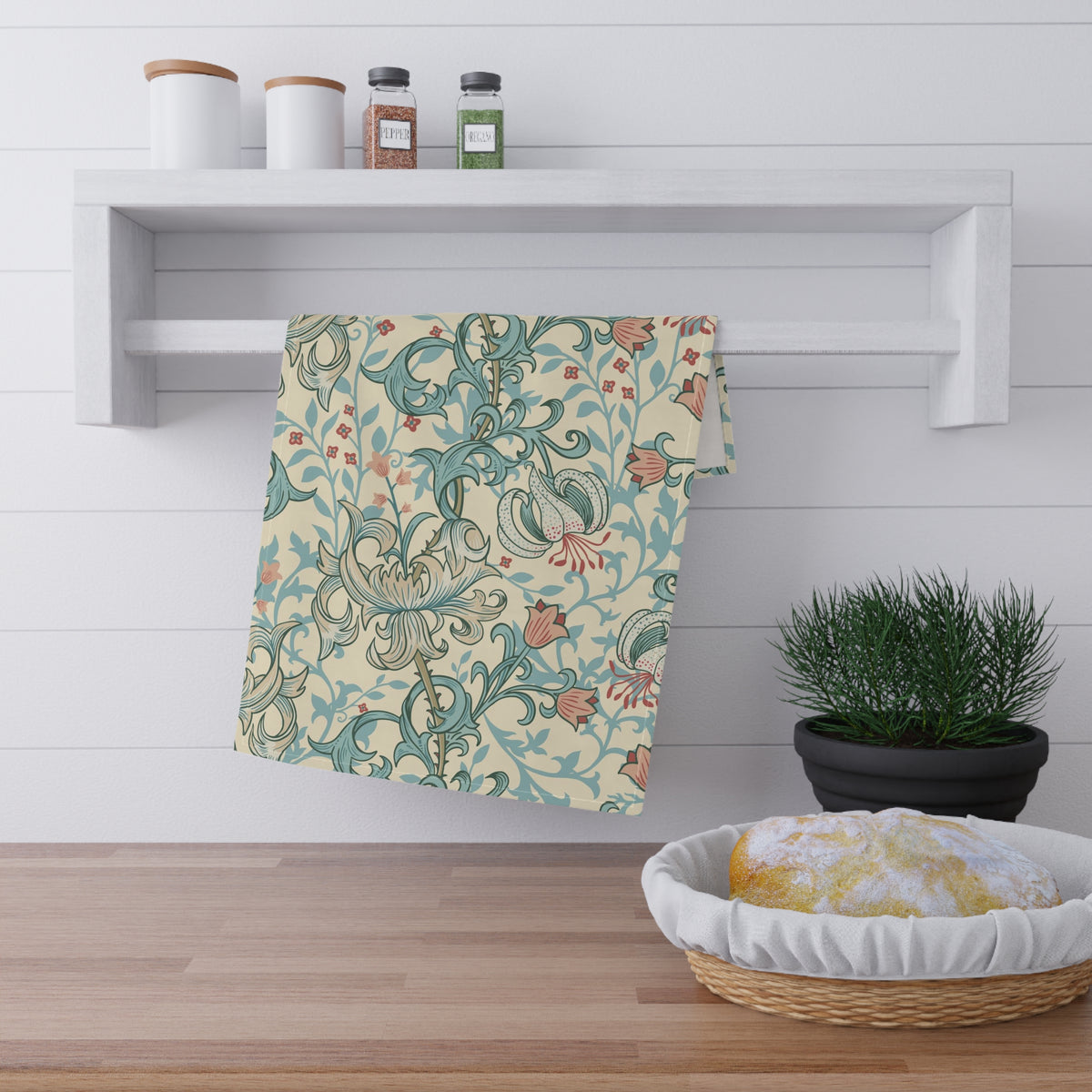 A patterned kitchen towel with a teal and rust botanical design on a cream background, hanging from a white wall-mounted rack in a bright kitchen setting.
