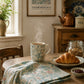 A styled breakfast scene in a rustic kitchen or dining area. A floral ceramic mug sits on a wooden table with steam rising, next to a croissant on a plate, a folded patterned cloth, a small bowl of jam, and a glass jar of granola.