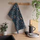 Decorative William Morris & Co tea towel with owl pattern hanging on a white shelf above a wooden surface with a basket and bread.