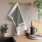 Tea towel with green pattern and text hanging on a wooden rack in a kitchen setting.