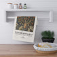 Decorative towel with a design and text hanging on a wall above a wooden shelf with a plant and basket.