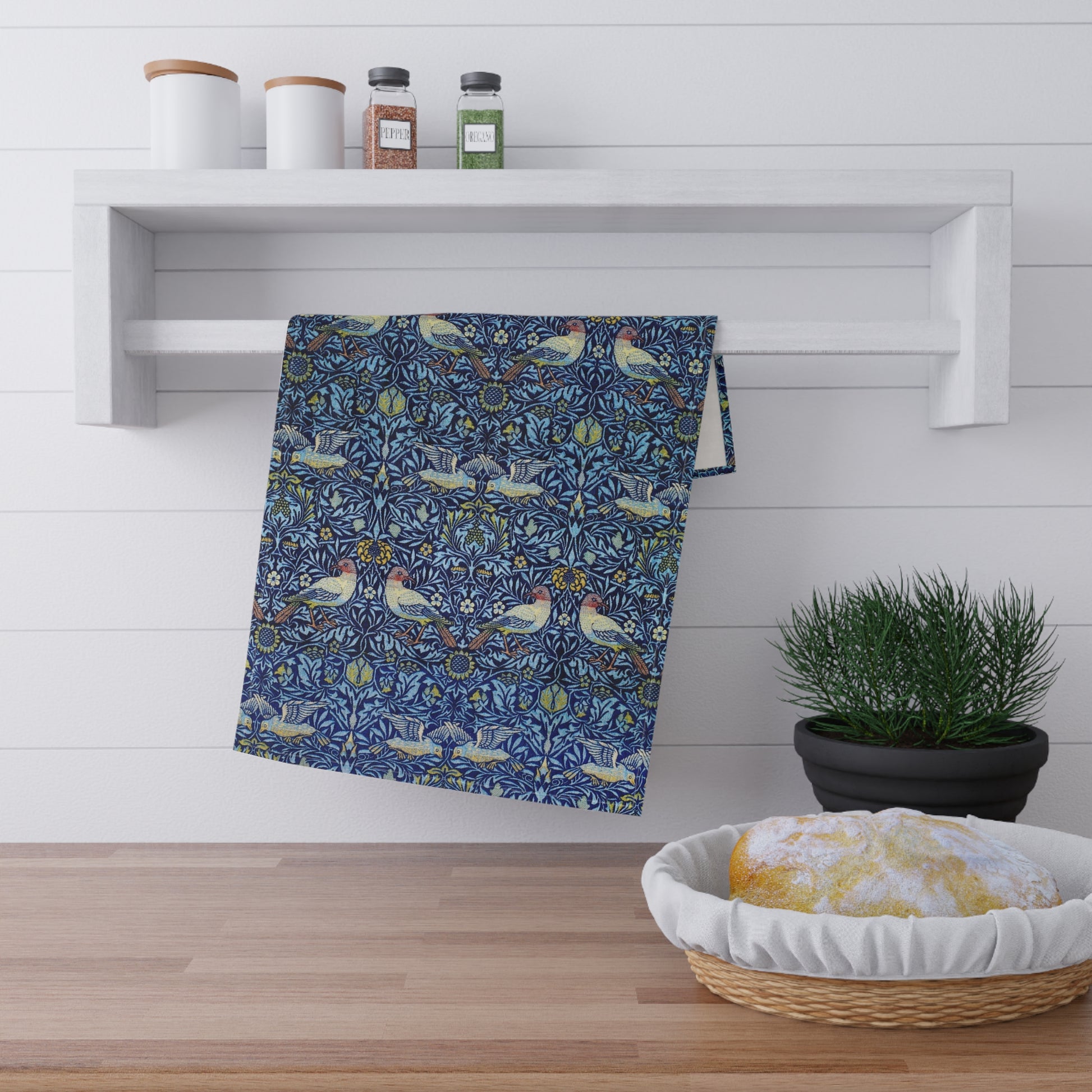 Decorative towel with bird pattern hanging on a white shelf above a wooden surface with a basket of bread and a plant.
