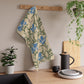 A kitchen tea towel with a William Morris & Co floral pattern featuring blue irises, birds, and a beige background.