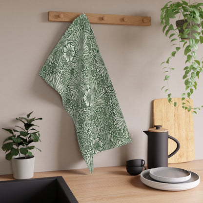 A kitchen tea towel with a floral and geometric pattern, hanging on a wooden hook on a wall, above a countertop with a plant and kitchen accessories.