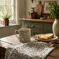 A ceramic mug with a green and pink botanical leaf pattern sits on a rustic wooden table next to a plate of toast and a matching patterned table runner. The scene is set in a cozy kitchen with a green cabinet, copper kettle, and a window in the background.
