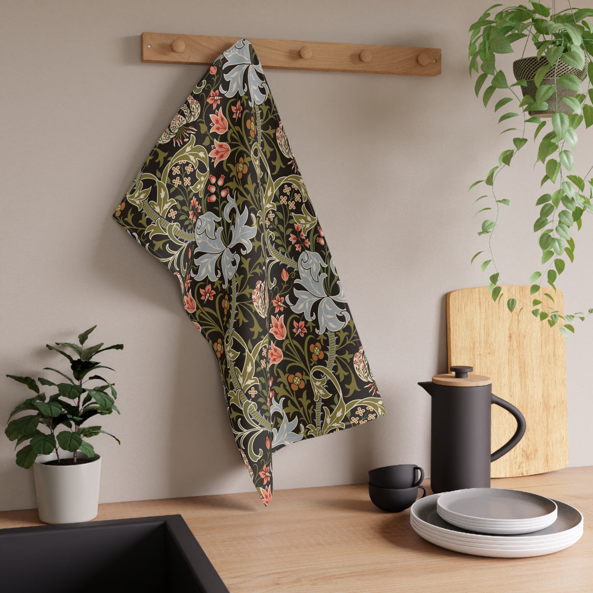 A kitchen towel with a William Morris-style floral pattern in muted green, pink, and gray hangs from a wooden peg on a beige wall above a wooden countertop with a black teapot, plates, and potted plants.