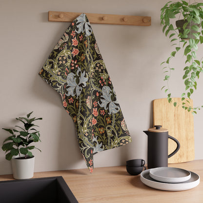A kitchen towel with a William Morris-style floral pattern in muted green, pink, and gray hangs from a wooden peg on a beige wall above a wooden countertop with a black teapot, plates, and potted plants.
