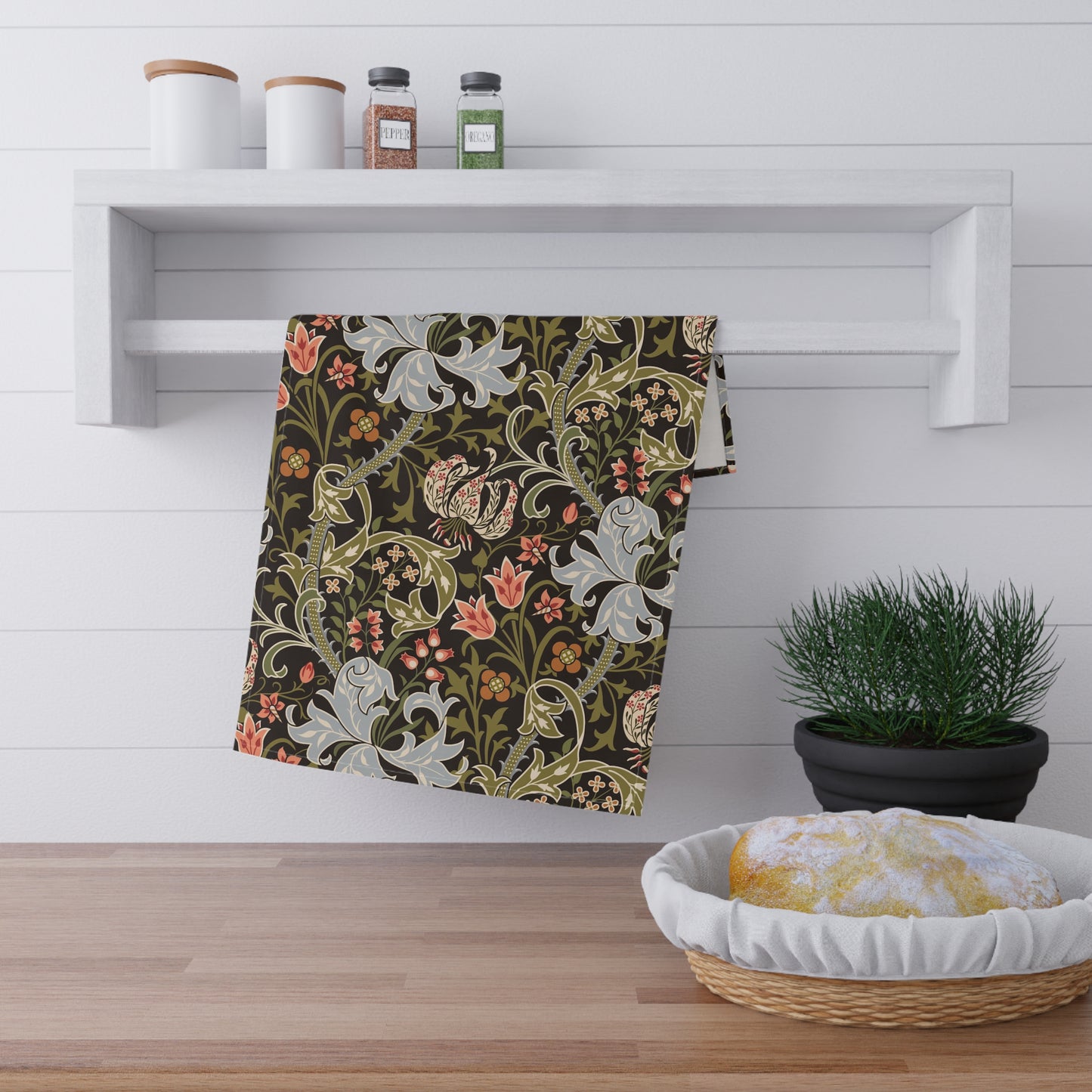 A decorative kitchen towel with a William Morris-style floral pattern in olive green, cream, and coral is hanging from a white wall-mounted rack in a bright kitchen setting.