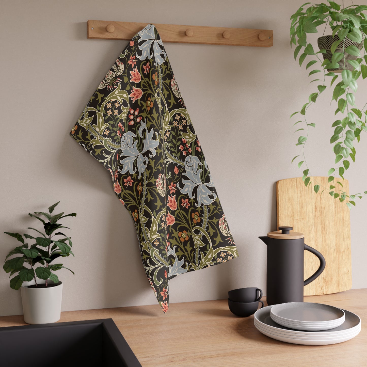 A kitchen towel with a William Morris-style floral pattern in muted green, pink, and gray hangs from a wooden peg on a beige wall above a wooden countertop with a black teapot, plates, and potted plants.