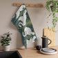 A kitchen tea towel featuring a repeated pattern of a white swan with a green and brownish background, and a set of dishes with cutlery on the side.
