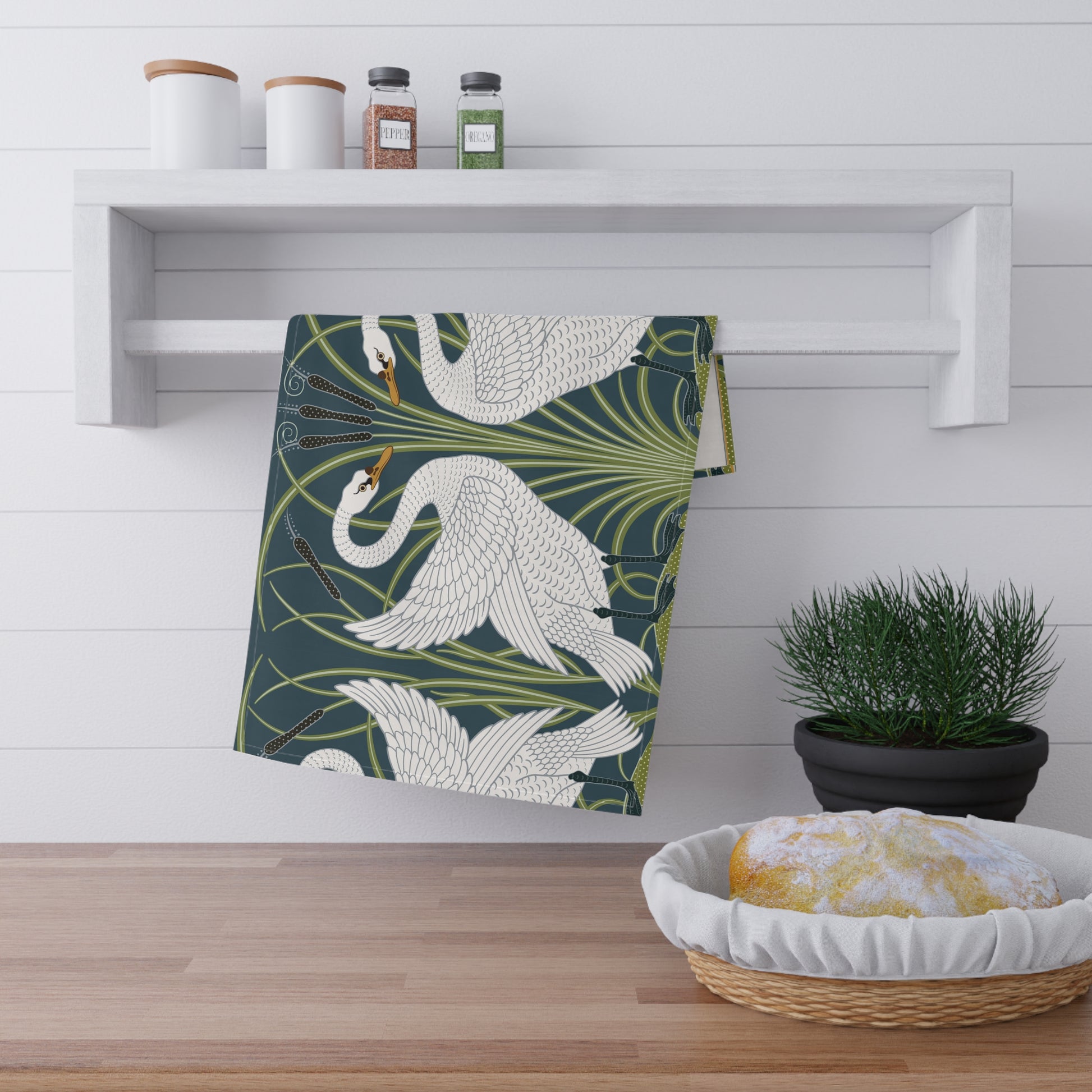 A kitchen tea towel featuring a repeated pattern of a white swan with a green and brownish background, and a set of dishes with cutlery on the side.