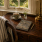 Steaming cup of tea on a wooden desk with a notebook and pen, next to a window with a view of greenery.