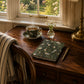 Teacup with saucer, notebook, and pen on a wooden desk by a window with floral view.
