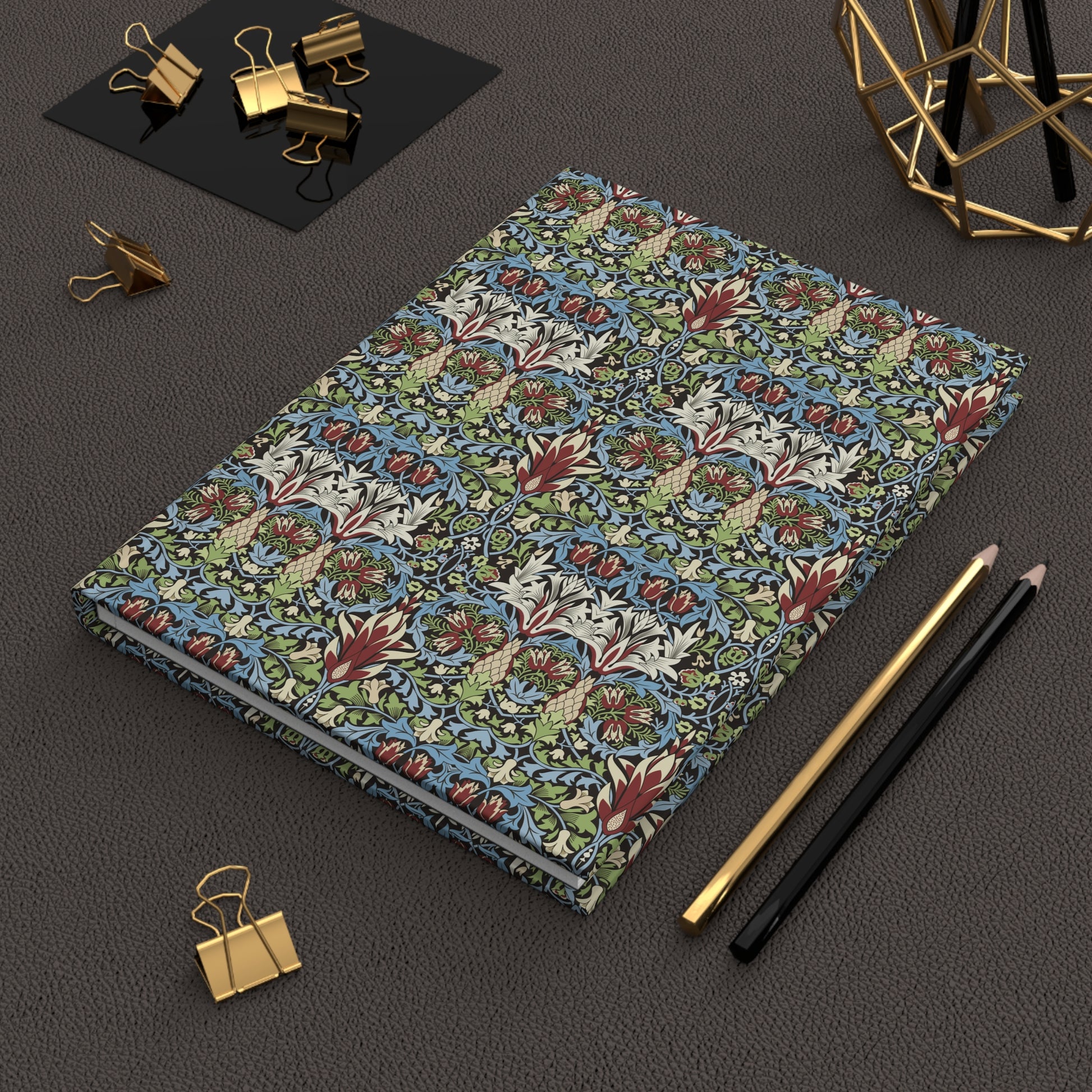 A hardcover notebook with a dense botanical and floral pattern in blue, green, red, and cream, styled on a dark textured surface with gold binder clips, pencils, and a geometric gold stand.