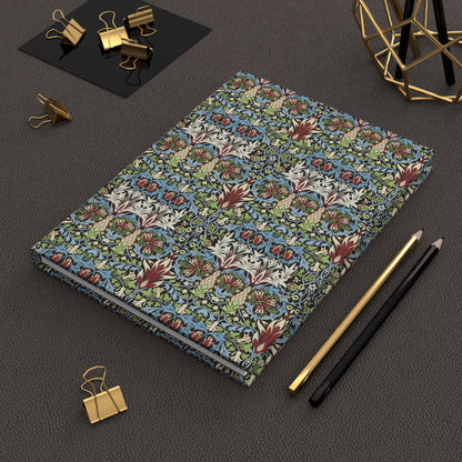 A hardcover notebook with a dense botanical and floral pattern in blue, green, red, and cream, styled on a dark textured surface with gold binder clips, pencils, and a geometric gold stand.