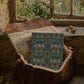 A brown leather bag with a cream sherpa lining sits open on a stone windowsill, with a patterned textile or book cover placed inside. The background shows a rustic stone wall and a window with outdoor greenery visible.