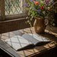 An open lined notebook lies on a rustic wooden table in warm sunlight, with a ceramic pitcher of wildflowers beside it and a window with diamond panes in the background.