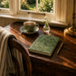 Tea cup with saucer, notebook, and pen on a wooden desk by a window with a view of greenery.