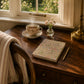 Tea cup with steam, notebook, pen, and small vase on a wooden table by a window with garden view.