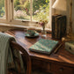 Wooden desk with a teacup, notebook, and books in a sunlit room.