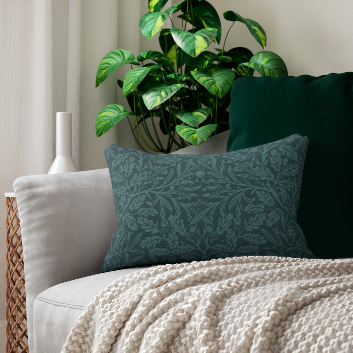 A dark teal patterned throw pillow resting on a light gray sofa, styled with a cream knit blanket, a dark green pillow, and a potted green plant in the background.
