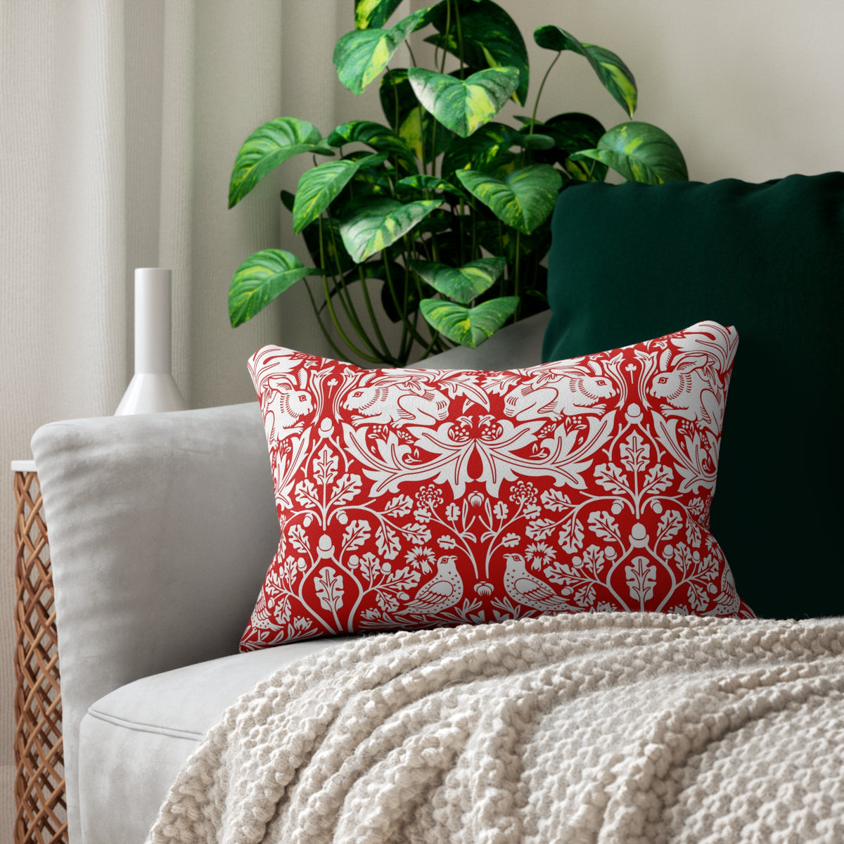 A red and white patterned decorative throw pillow displayed on a light gray sofa with a cream knit blanket, next to a dark green pillow, with a potted green plant in the background.