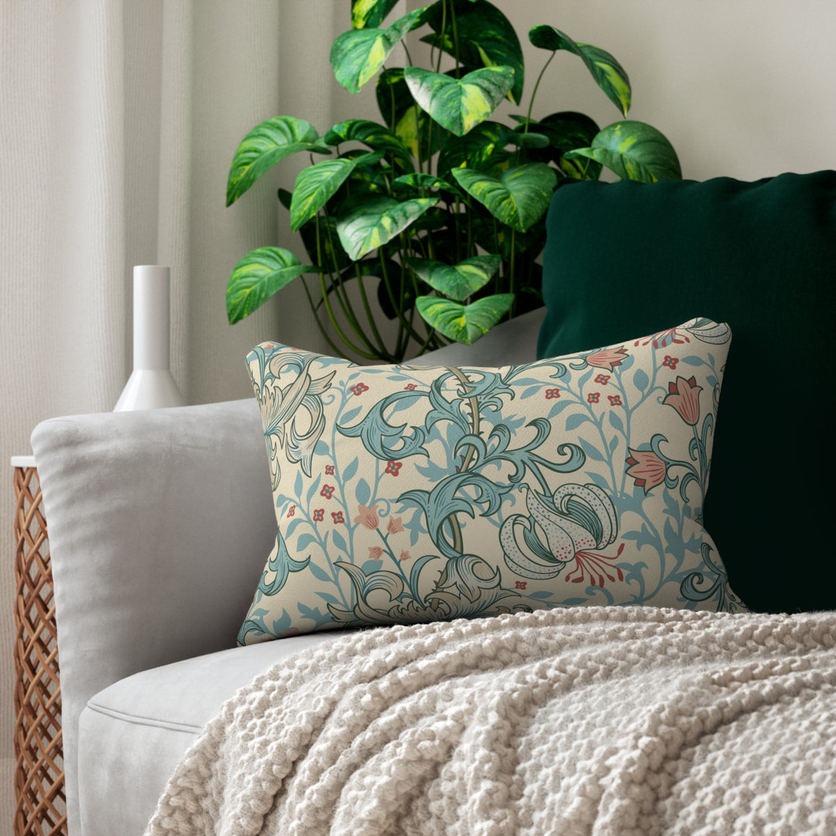 A decorative throw pillow with a vintage floral and scroll pattern in teal, pink, and cream, styled on a light gray sofa next to a dark green pillow and a textured cream knit blanket.