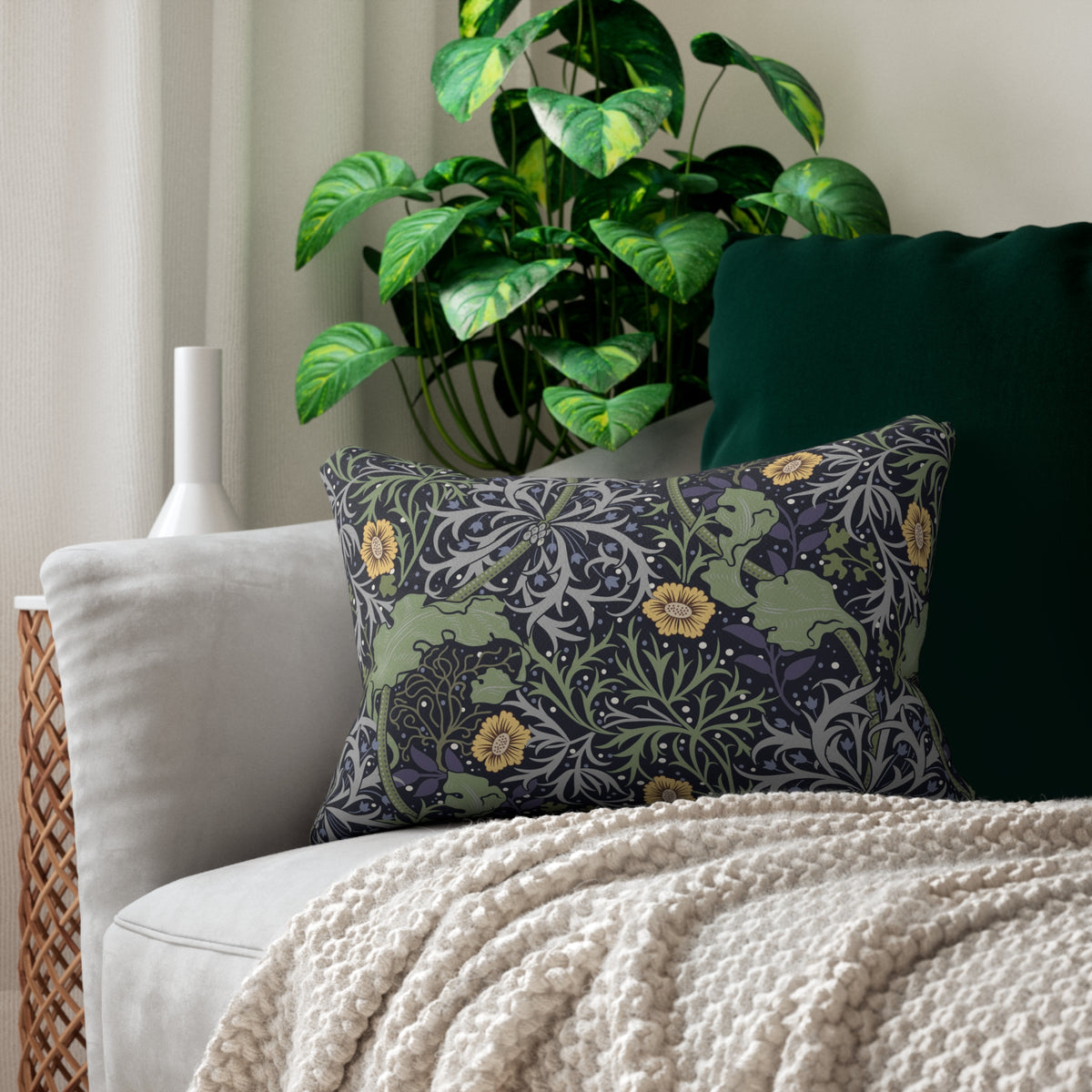 A decorative throw pillow with a dark floral and botanical pattern sits on a light gray sofa, styled with a cream knit blanket and a dark green cushion, with a potted green plant in the background.