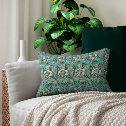 A decorative rectangular throw pillow with a teal and white floral pattern, styled on a light gray sofa with a cream knit blanket and a dark green cushion in a living room setting.