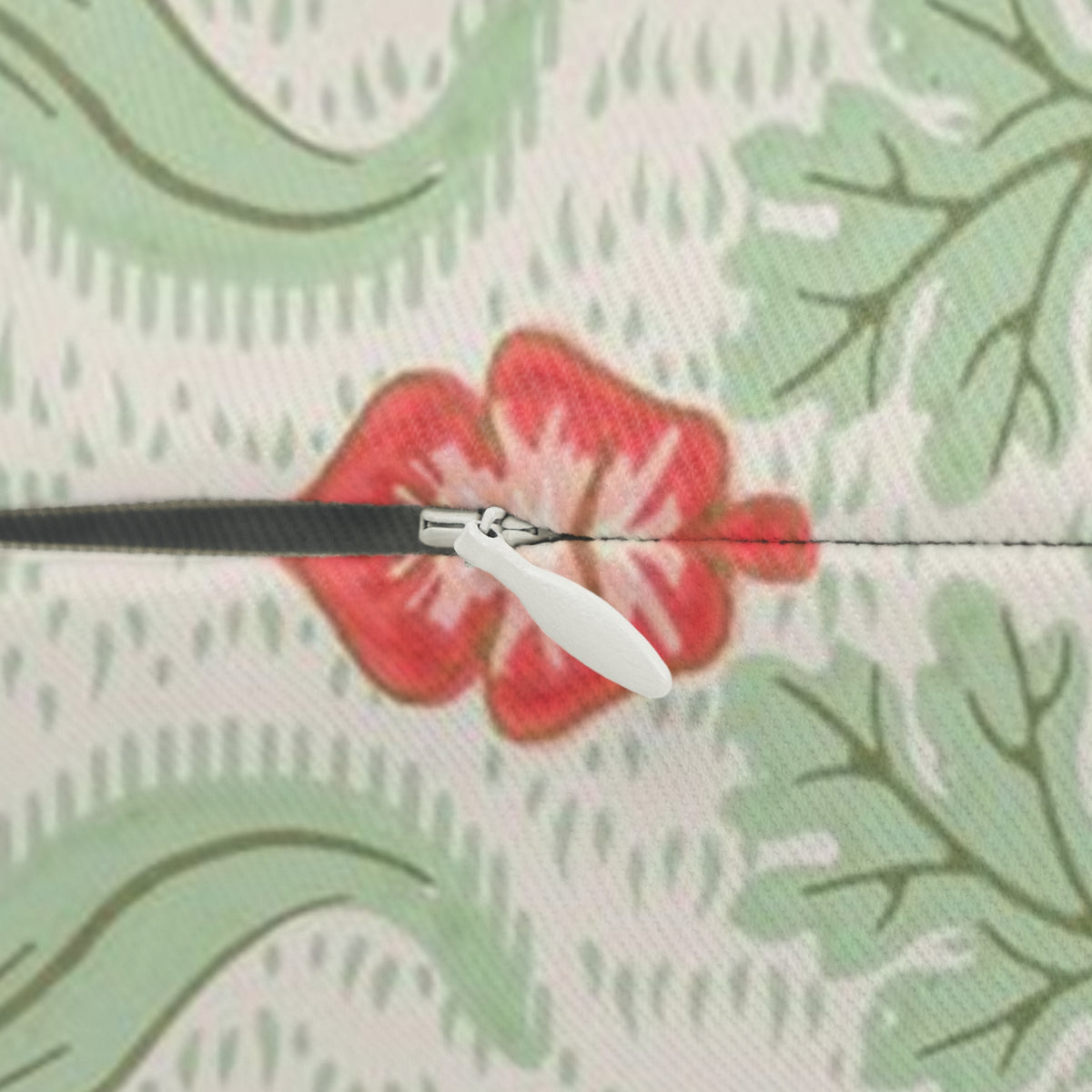 A close-up of a zipper with a white pull tab and black teeth, set against a patterned fabric background featuring light green leaves and a red floral motif.