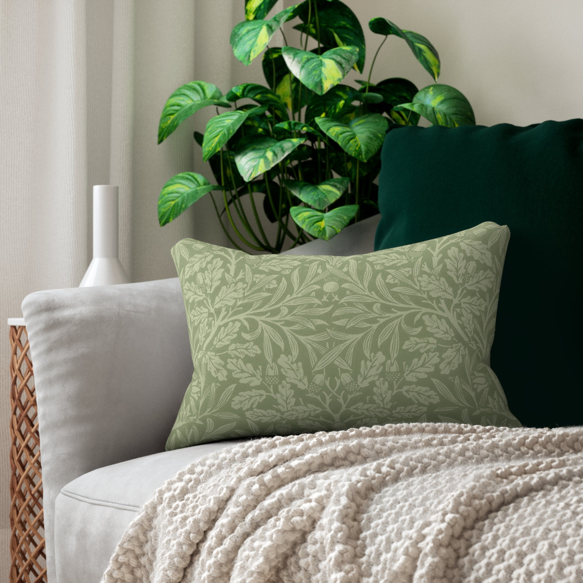 A sage green decorative throw pillow with a light green damask leaf pattern, styled on a light gray sofa with a cream knit blanket and a dark green pillow behind it.