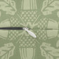 Close-up of a zipper with a white pull tab and black teeth, set against a sage green fabric background with a light cream abstract pattern.